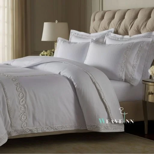 A hotel bed with white sheet & pillows, comforter having embroidery on borders.